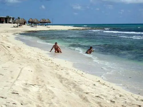 Two people wade in shallow turquoise water off a sandy beach with thatched-roof structures and palm trees along the shore