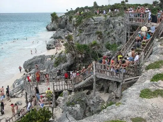 Tourists navigate wooden stairways and platforms built into rocky cliffs above a sandy beach and turquoise water