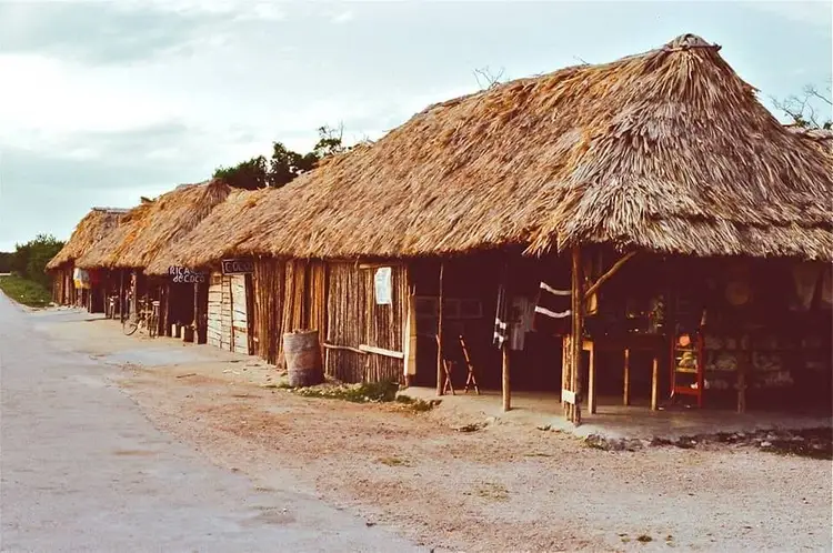 Row of thatched-roof structures with wooden walls line a dirt path in a rural coastal area