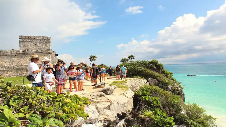 Tour group stands near a stone tower overlooking turquoise water and tropical vegetation on a clifftop archaeological site