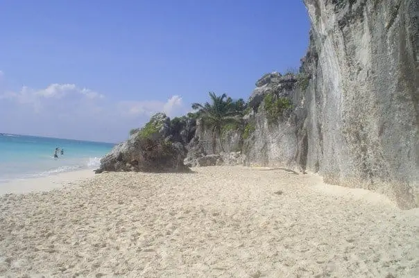 Sandy beach with tall limestone cliffs, tropical vegetation, and turquoise water where a few swimmers wade near shore