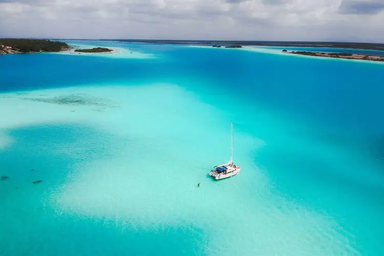 Sailboat anchored in shallow turquoise water near small islands and sandy shores under a cloudy sky