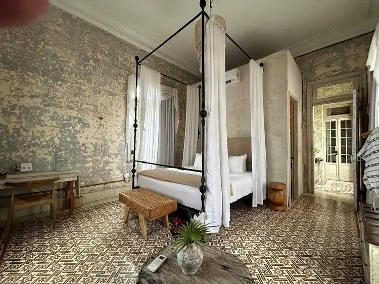 A four-poster bed with white curtains sits in a hotel bedroom with aged wallpaper, patterned tile flooring, and wooden furnishings