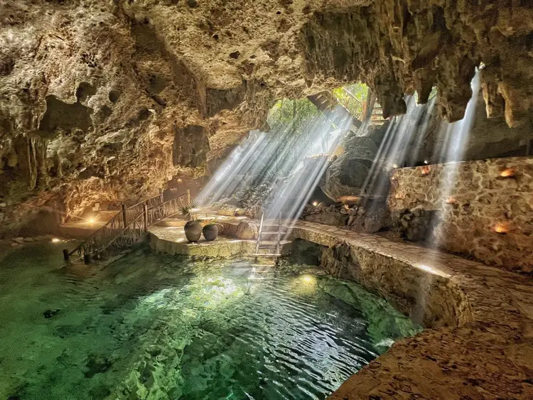 Sunlight streams through openings in a limestone cave above an underground pool with stone walkways and railings