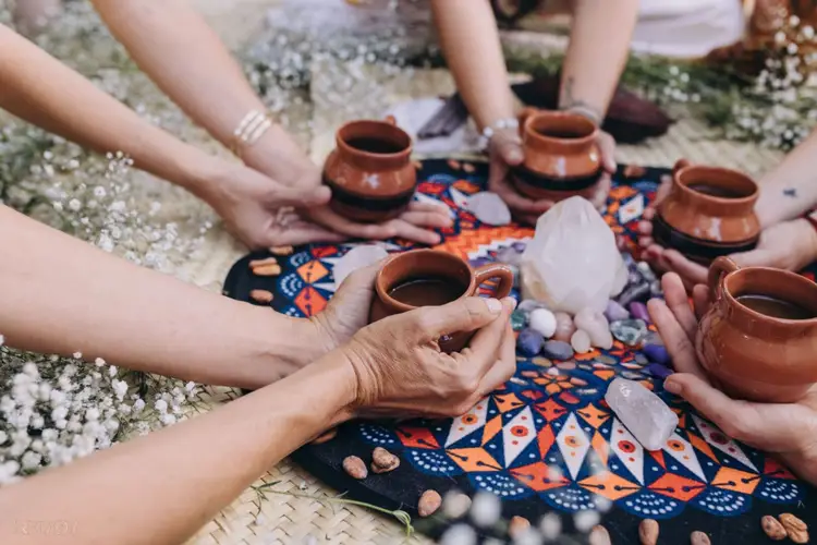 Multiple people sit in a circle holding clay cups and passing items around a patterned textile surrounded by white flowers