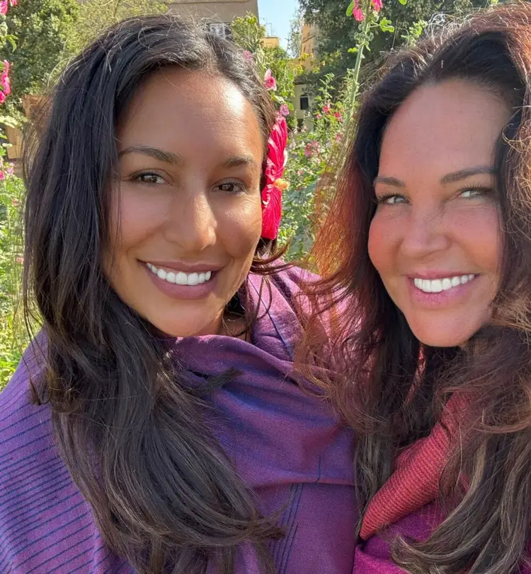 Two women smile at the camera in front of a vine-covered wall with pink flowers and green foliage in sunlight