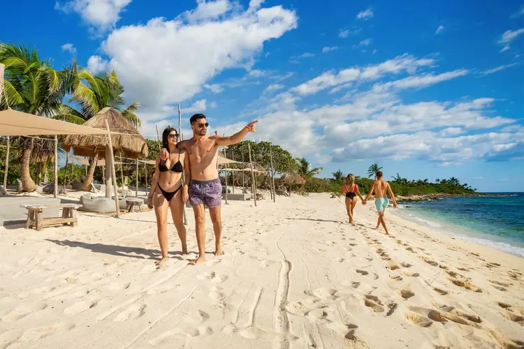 A couple stands on a sandy beach while the man points toward the water, with other beachgoers visible in the distance and thatched umbrellas lining the shore