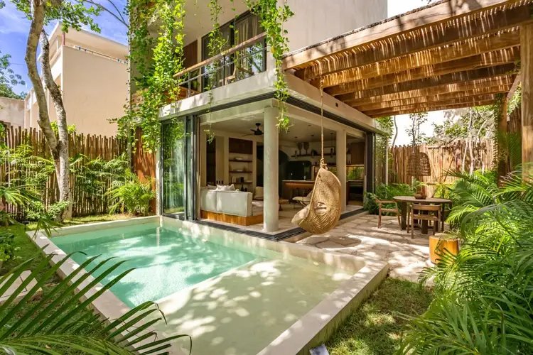 Modern home with pool and tropical landscaping features floor-to-ceiling windows opening to a patio with lounge chairs and hanging egg chair Modern home with pool and tropical landscaping features floor-to-ceiling windows opening to a patio with lounge chairs and hanging egg chair