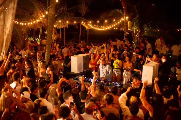 A DJ performs for a large crowd dancing at an outdoor nighttime event under string lights and palm trees A DJ performs for a large crowd dancing at an outdoor nighttime event under string lights and palm trees
