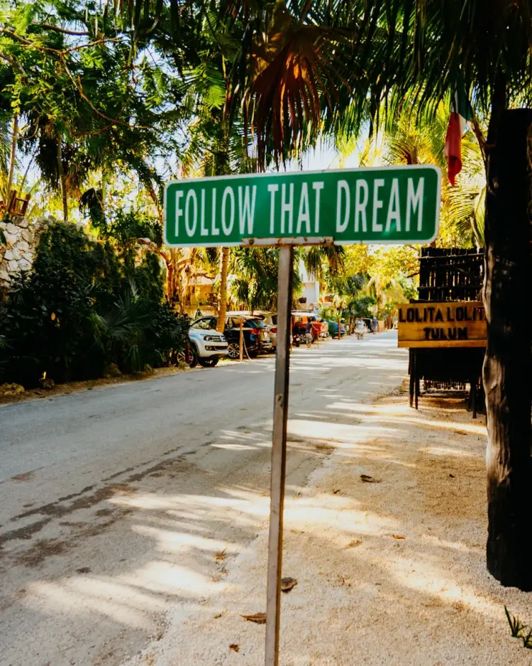 A green street sign reading "Follow That Dream" stands on a sandy road lined with palm trees and tropical vegetation A green street sign reading "Follow That Dream" stands on a sandy road lined with palm trees and tropical vegetation