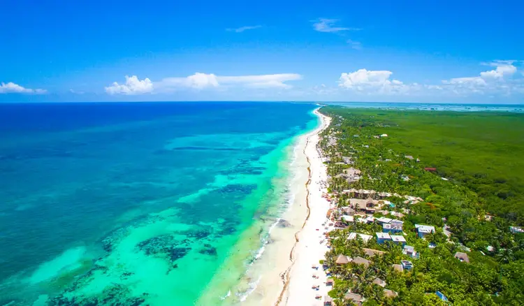Aerial view of a curved white sand beach meeting turquoise water, with dense green forest and scattered buildings along the shoreline under blue sky with white Aerial view of a curved white sand beach meeting turquoise water, with dense green forest and scattered buildings along the shoreline under blue sky with white