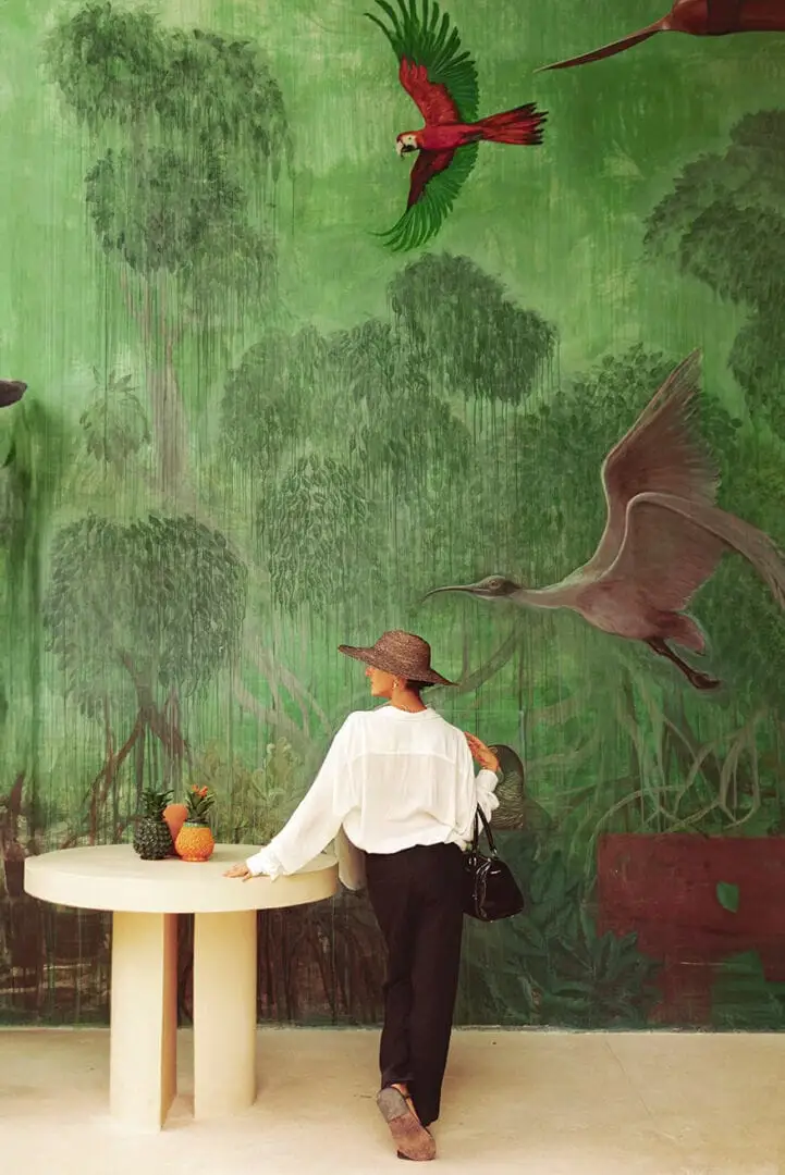 Person wearing a straw hat standing at a round table facing a large mural depicting wetland vegetation and flying birds Person wearing a straw hat standing at a round table facing a large mural depicting wetland vegetation and flying birds