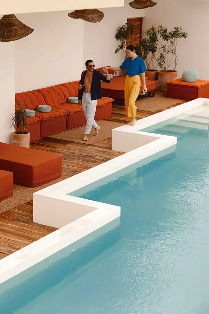 Two people stand in a modern lounge area next to an indoor pool, surrounded by orange seating and woven ceiling panels Two people stand in a modern lounge area next to an indoor pool, surrounded by orange seating and woven ceiling panels