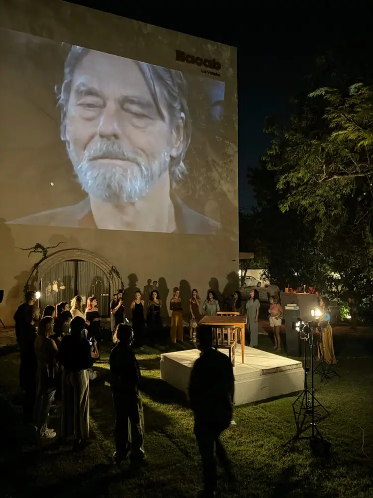 People gather for a nighttime outdoor event with a large projected portrait displayed on a wall behind them People gather for a nighttime outdoor event with a large projected portrait displayed on a wall behind them
