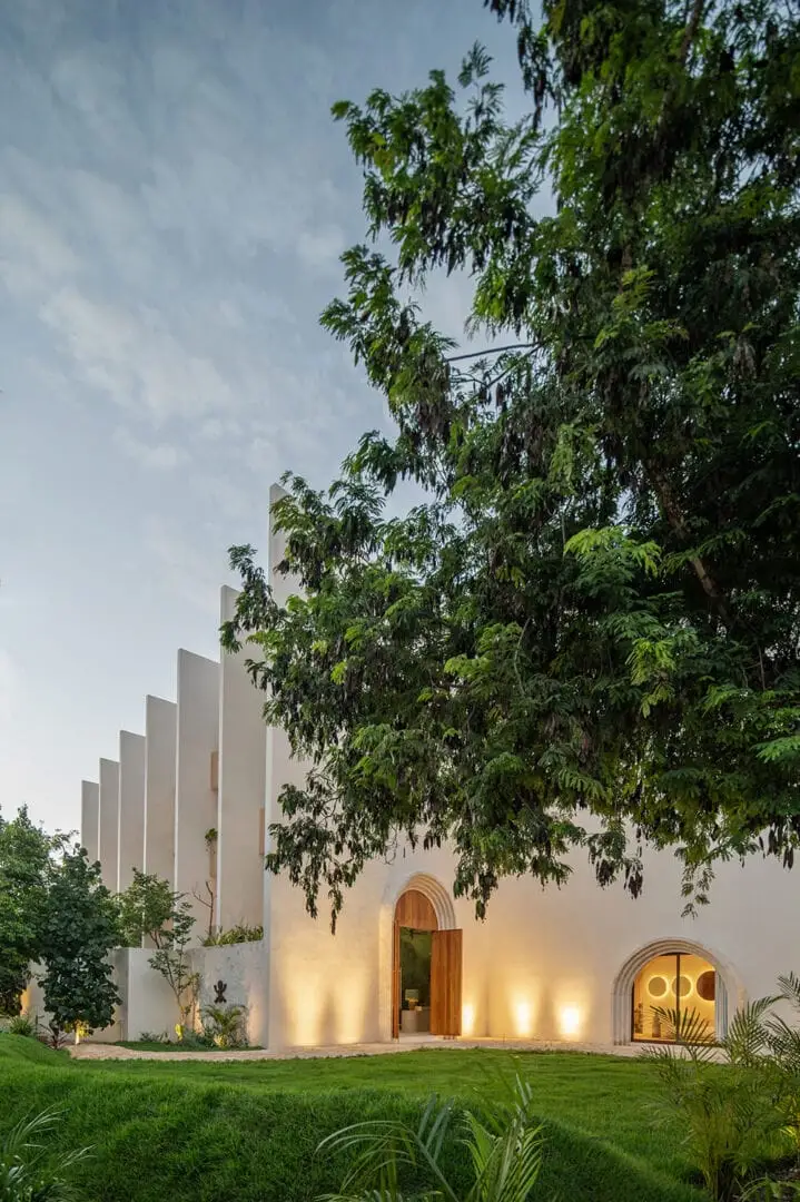 A modern white building with vertical ribbed panels and arched wooden doors sits behind a lawn with overhanging tree branches in evening light A modern white building with vertical ribbed panels and arched wooden doors sits behind a lawn with overhanging tree branches in evening light