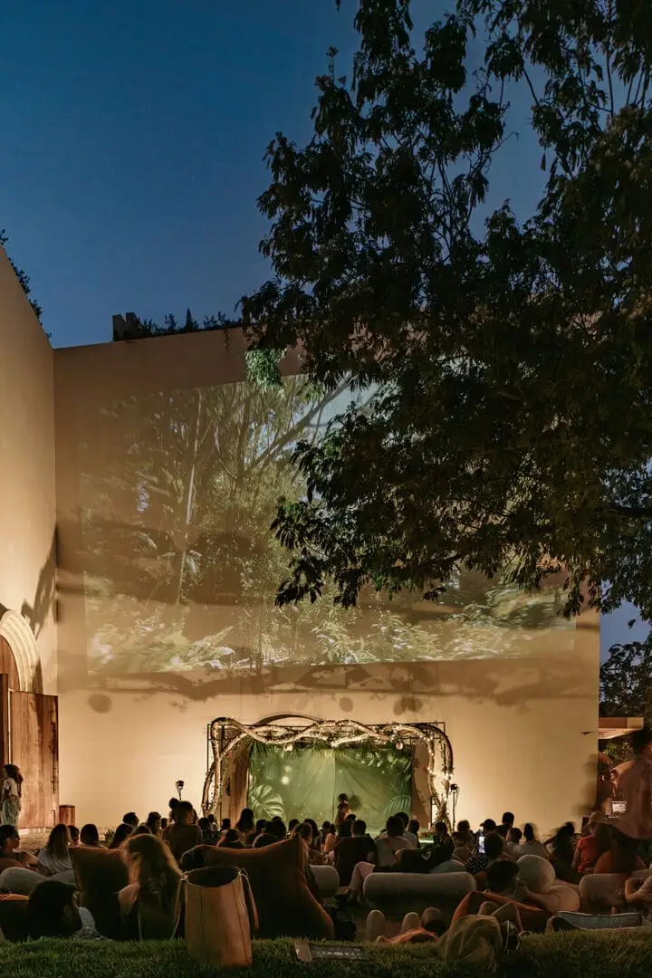 Crowd seated on grass watches a performance on an illuminated outdoor stage beneath trees at dusk Crowd seated on grass watches a performance on an illuminated outdoor stage beneath trees at dusk