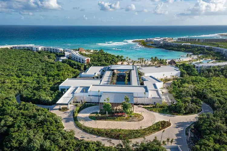 Aerial view of a modern white resort building surrounded by dense green vegetation, with a turquoise ocean and beach visible beyond Aerial view of a modern white resort building surrounded by dense green vegetation, with a turquoise ocean and beach visible beyond