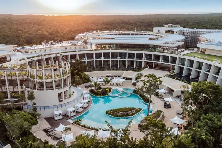Large resort complex with curved white buildings surrounding a blue pool with island landscaping, surrounded by forested terrain Large resort complex with curved white buildings surrounding a blue pool with island landscaping, surrounded by forested terrain