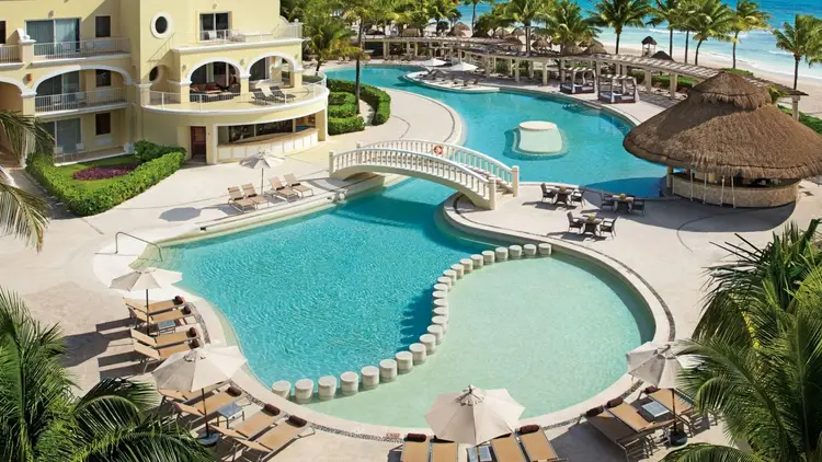 Aerial view of a beachfront resort with multiple connected swimming pools, lounge chairs, a thatched-roof palapa, and a yellow curved building on a tropical coa Aerial view of a beachfront resort with multiple connected swimming pools, lounge chairs, a thatched-roof palapa, and a yellow curved building on a tropical coa