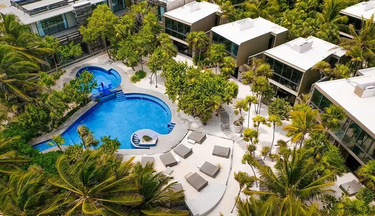 Aerial view of a resort with an irregularly shaped blue swimming pool surrounded by lounge chairs, palm trees, and modern buildings with white roofs Aerial view of a resort with an irregularly shaped blue swimming pool surrounded by lounge chairs, palm trees, and modern buildings with white roofs