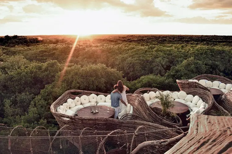 Person standing on an elevated deck with woven seating areas overlooking a forested landscape at sunset Person standing on an elevated deck with woven seating areas overlooking a forested landscape at sunset