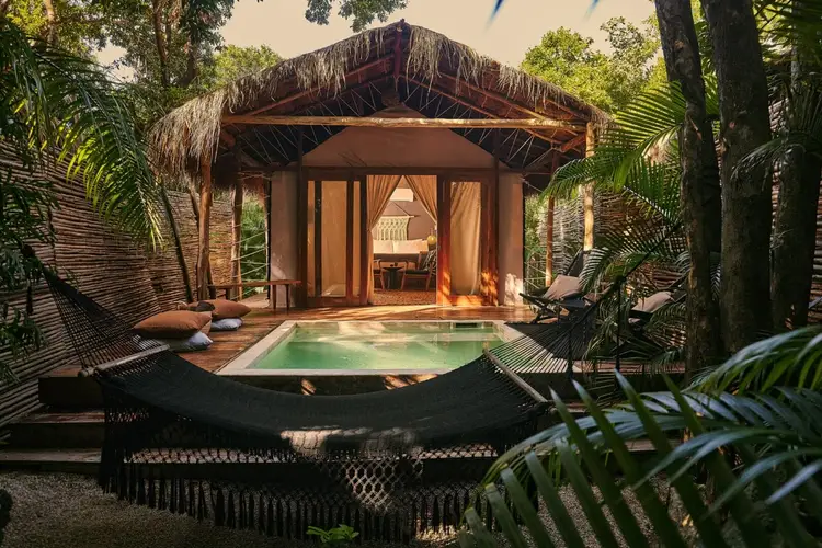 A thatched-roof bungalow with an outdoor pool surrounded by hammocks and tropical vegetation in a forest setting A thatched-roof bungalow with an outdoor pool surrounded by hammocks and tropical vegetation in a forest setting