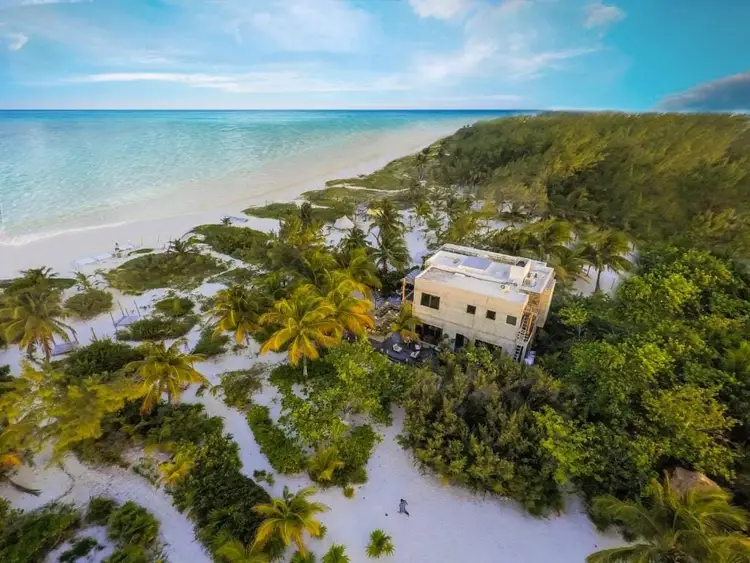 A beige building with solar panels sits among palm trees and vegetation on white sand near turquoise shallow water and beach A beige building with solar panels sits among palm trees and vegetation on white sand near turquoise shallow water and beach