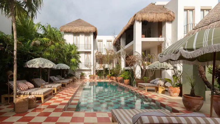 A resort courtyard features a rectangular pool surrounded by thatched-roof bungalows, striped loungers, umbrellas, potted plants, and a red and white checkered A resort courtyard features a rectangular pool surrounded by thatched-roof bungalows, striped loungers, umbrellas, potted plants, and a red and white checkered