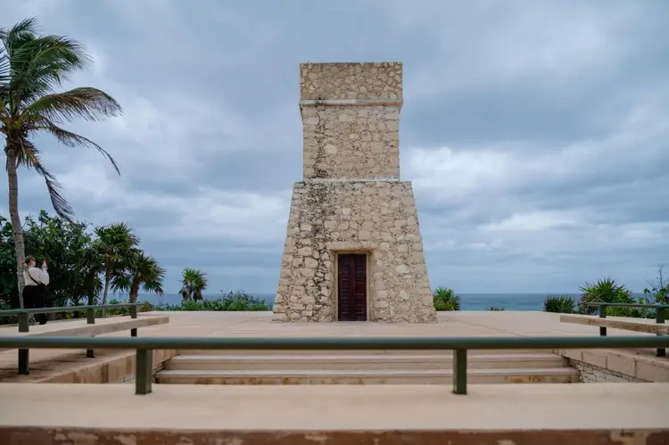 Stone tower structure with a wooden door surrounded by a paved plaza with benches, palm trees, and ocean view in background