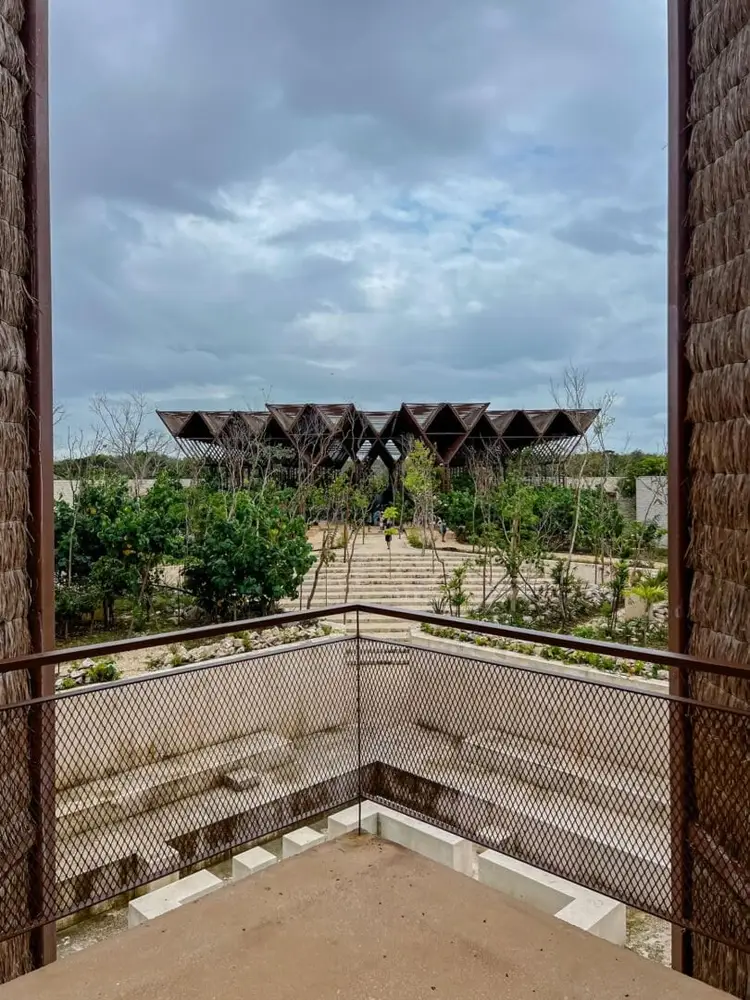 View from a balcony overlooking a modern geometric building with stepped architecture surrounded by planted gardens and trees under a cloudy sky