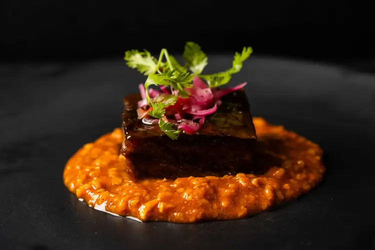 A dark rectangular piece of protein sits atop an orange puree, garnished with microgreens and pink pickled vegetables on a black plate A dark rectangular piece of protein sits atop an orange puree, garnished with microgreens and pink pickled vegetables on a black plate