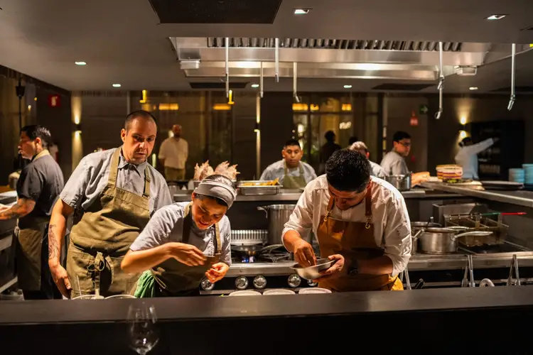Chefs work at a stovetop in an open kitchen while other staff members move about in the background Chefs work at a stovetop in an open kitchen while other staff members move about in the background