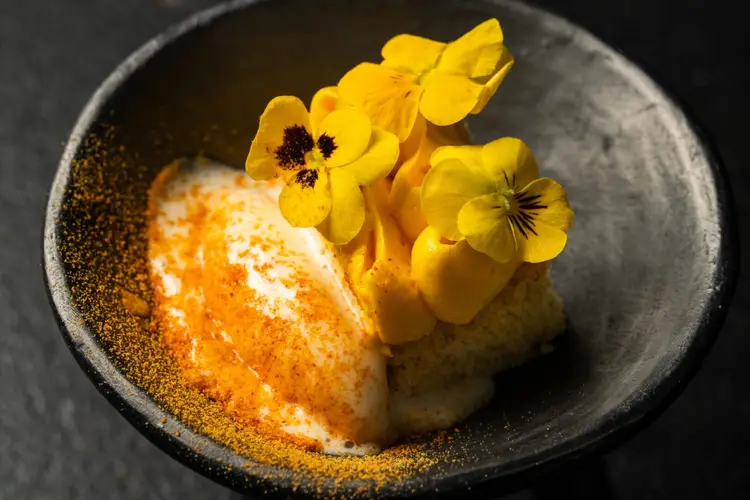 A plated dessert with a creamy center dusted with golden powder, garnished with bright yellow pansies in a dark ceramic bowl A plated dessert with a creamy center dusted with golden powder, garnished with bright yellow pansies in a dark ceramic bowl