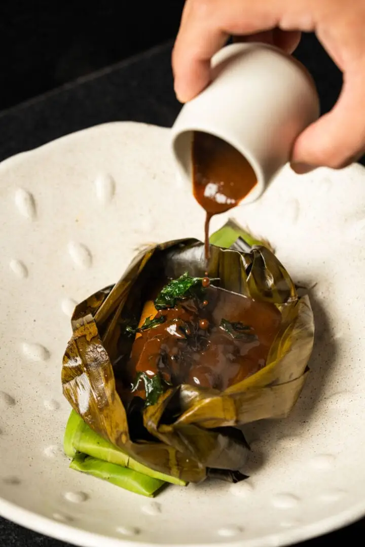 Hand pouring brown sauce over a leaf-wrapped dumpling garnished with greens on a white plate Hand pouring brown sauce over a leaf-wrapped dumpling garnished with greens on a white plate