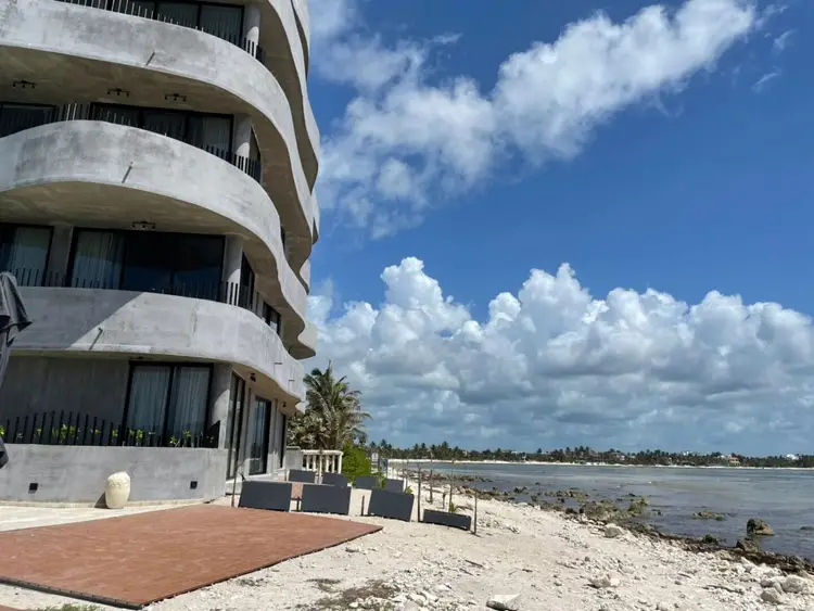 Curved concrete building with multiple balconies sits on a sandy beach next to shallow water under a blue sky with white clouds Curved concrete building with multiple balconies sits on a sandy beach next to shallow water under a blue sky with white clouds