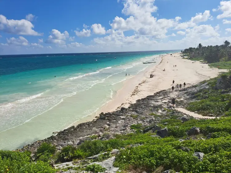 Wide sandy beach with turquoise water and small waves, scattered visitors in the water and on shore, rocky outcrops and tropical vegetation in the foreground, p
