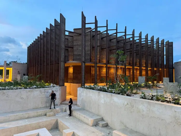 Modern building with dark metal vertical slats and illuminated orange interior walls, surrounded by concrete terraces where two people stand near the entrance