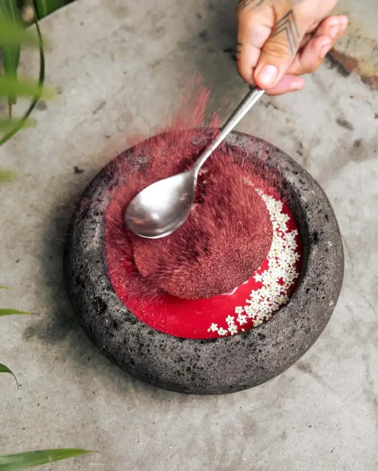 Hand holding a spoon over a stone bowl containing red paste garnished with white seeds on concrete ground Hand holding a spoon over a stone bowl containing red paste garnished with white seeds on concrete ground