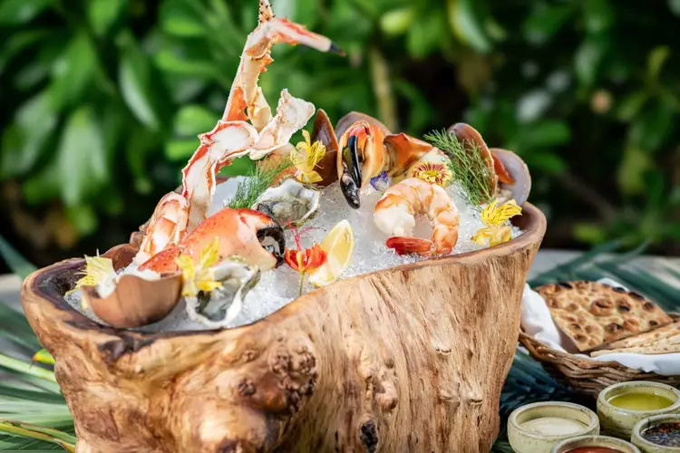 Cooked shrimp garnished with flowers and microgreens arranged in a wooden bowl surrounded by green foliage and condiment cups Cooked shrimp garnished with flowers and microgreens arranged in a wooden bowl surrounded by green foliage and condiment cups