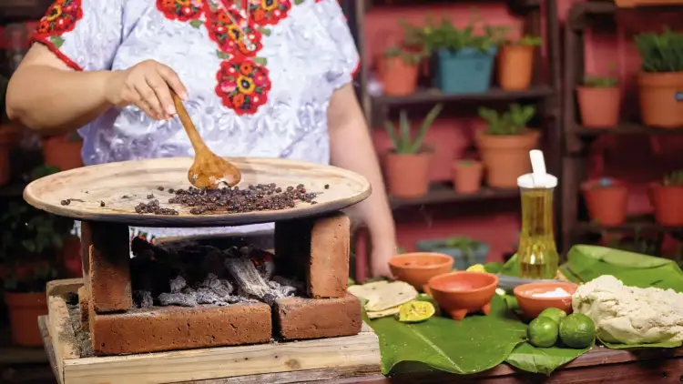 Person in embroidered white shirt roasts coffee beans on a clay comal over a brick stove, with potted plants and ingredients visible in background Person in embroidered white shirt roasts coffee beans on a clay comal over a brick stove, with potted plants and ingredients visible in background