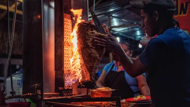 A vendor cuts meat from a vertical spit at a street food stand, with flames and heat visible in the background A vendor cuts meat from a vertical spit at a street food stand, with flames and heat visible in the background