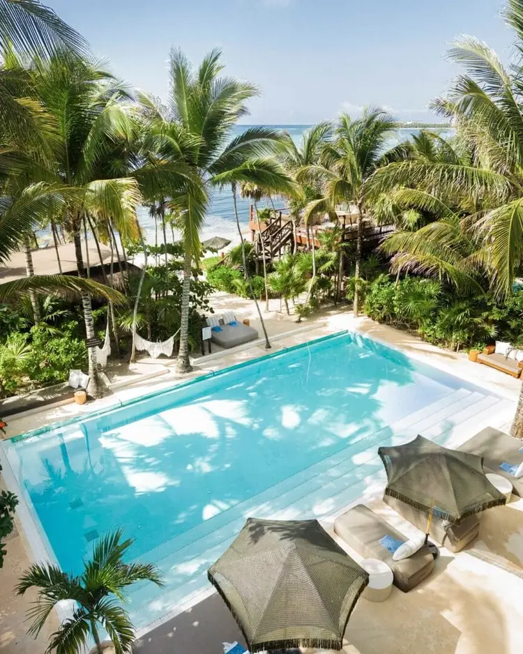A rectangular swimming pool with turquoise water sits surrounded by palm trees, lounge chairs with umbrellas, and a beach with wooden structures visible beyond