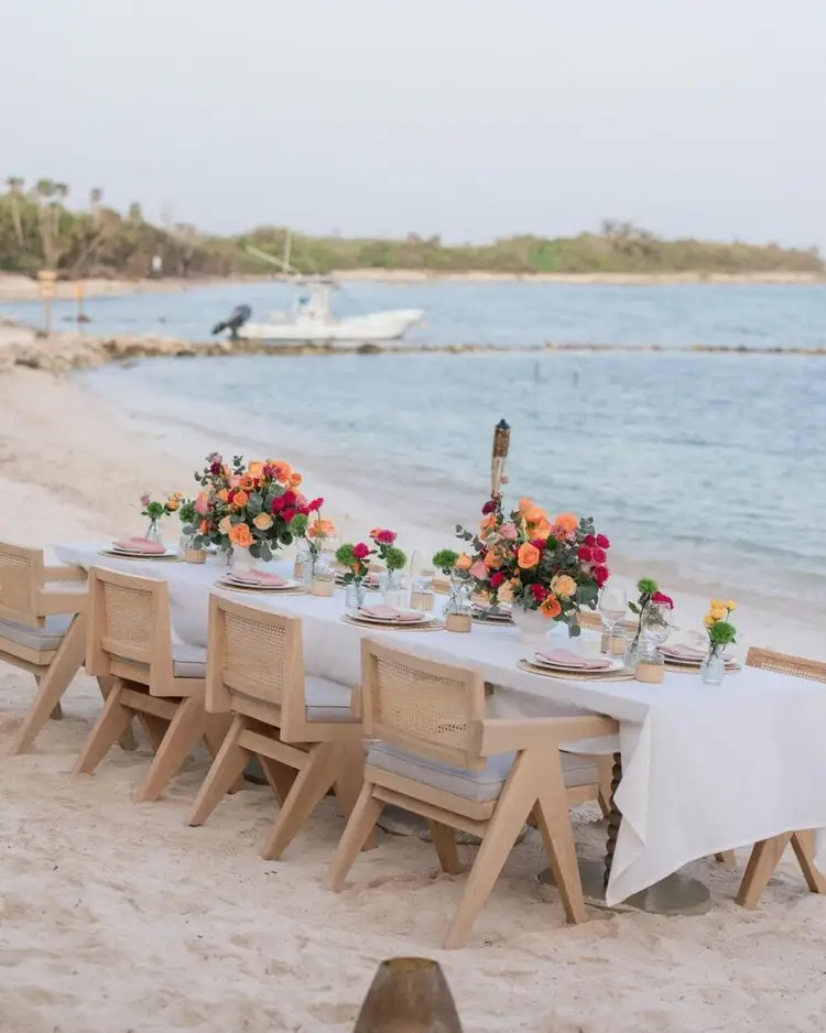 A long table set with place settings and colorful floral arrangements sits on a sandy beach overlooking calm water with boats and mangrove trees in the distance
