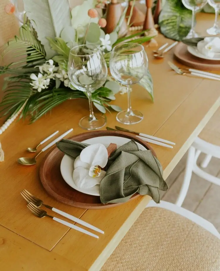 A formally set dining table with wooden placemats, white plates on brown chargers, green napkins, wine glasses, and a central floral arrangement of green leaves
