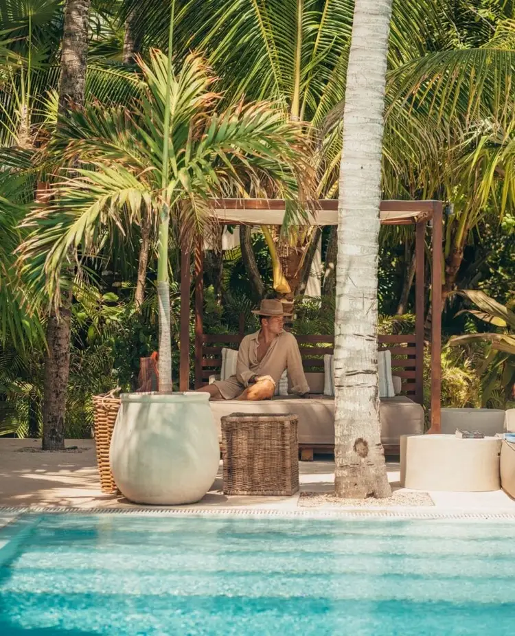 A person sits on a cushioned lounge surrounded by potted plants and palm trees beside a swimming pool