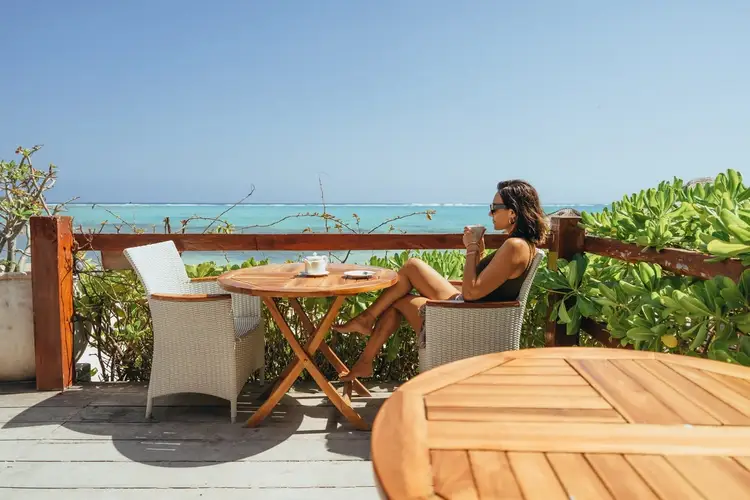 Woman sitting at a wooden table on a beachfront deck overlooking turquoise water and tropical vegetation