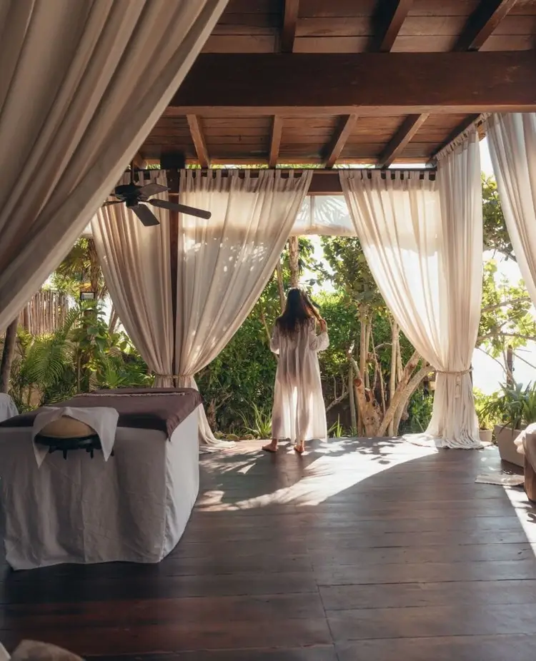 Person standing on a wooden deck under a pergola with flowing white curtains, surrounded by lush tropical vegetation
