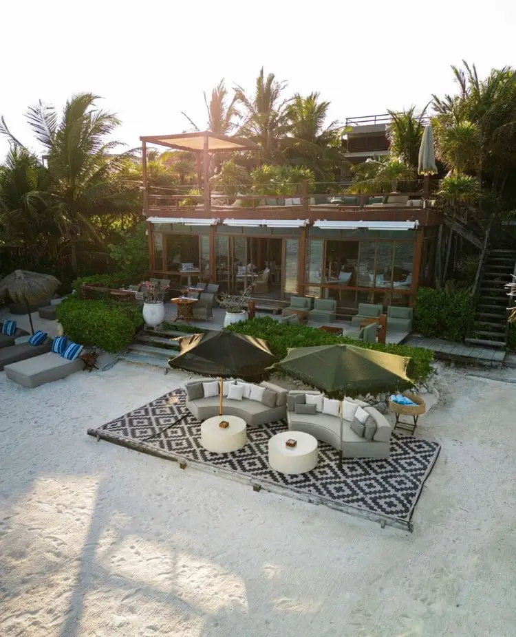 Beachfront resort with lounge seating under umbrellas on sand, modern glass building with upper deck, and palm trees surrounding the property