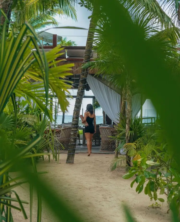 Woman in black dress walking on sandy path lined with palm trees toward a beachfront structure with white curtains and ocean view