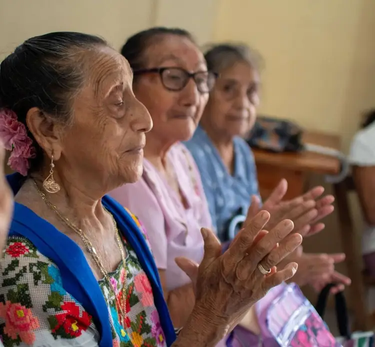 An older woman in a floral embroidered shirt claps her hands while sitting with two other women in an indoor setting with a pale yellow wall An older woman in a floral embroidered shirt claps her hands while sitting with two other women in an indoor setting with a pale yellow wall